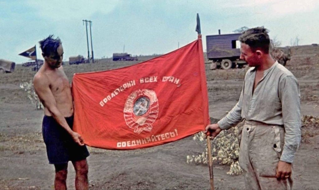 Two men on the steppe display a captured red Soviet flag beside field vehicles